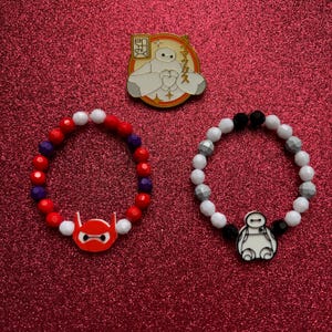Op de afbeelding: Twee kralenarmbanden met bedels met het personage Baymax uit de Disney-film Big Hero 6. De ene armband is rood, wit en paars, de andere is wit, grijs en zwart.