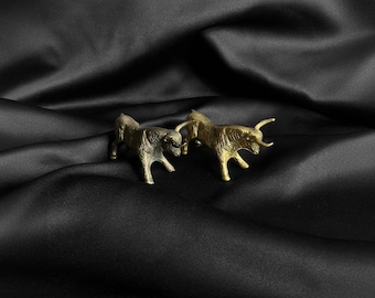 May include: Two gold-colored metal bull figurines facing each other on a black satin background.