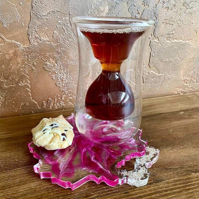 K&ouml;nnte beinhalten: Eine klare Glas-Teetasse in Sanduhrform mit einem Untersetzer aus rosa und wei&szlig;em Harz. Die Teetasse ist mit dunkelbraunem Tee gef&uuml;llt. Ein wei&szlig;er Keks mit Schokoladenst&uuml;ckchen liegt auf dem Untersetzer.
