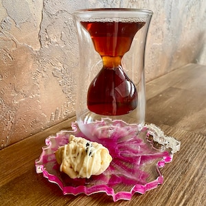 K&ouml;nnte beinhalten: Eine gl&auml;serne Teetasse in Sanduhrform mit einem Untersetzer aus rosa und wei&szlig;em Harz und einem wei&szlig;en Keks mit Schokoladenst&uuml;ckchen darauf.