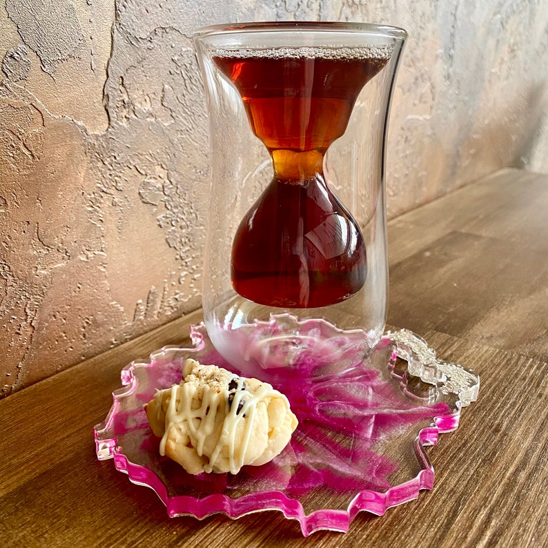 K&ouml;nnte beinhalten: Eine gl&auml;serne Teetasse in Sanduhrform mit einem Untersetzer aus rosa und wei&szlig;em Harz und einem wei&szlig;en Keks mit Schokoladenst&uuml;ckchen darauf.