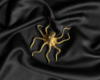 Puede incluir: Una figura de pulpo dorada con ocho tentáculos, sobre una tela negra y sedosa. El pulpo tiene una superficie texturizada, y la tela presenta pliegues suaves.