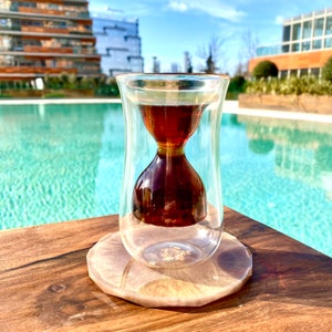 K&ouml;nnte beinhalten: Eine klare Glas-Teetasse in Sanduhrform mit braunem Tee darin, die auf einem beigefarbenen Untersetzer auf einer Holzoberfl&auml;che steht. Die Teetasse steht vor einem Schwimmbad.