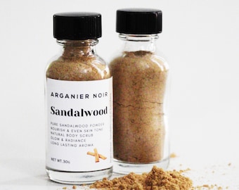May include: Two glass jars with black lids filled with a light brown powder. The label on the front jar reads "Arganier Noir Sandalwood Pure Sandalwood Powder Nourish & Even Skin Tone Natural Body Scrub Glow & Radiance Long Lasting Aroma Net Wt. 30g".