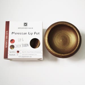 Puede incluir: Un pequeño tarro de terracota lleno de una mancha para labios y mejillas de color rojo-marrón. El tarro está etiquetado como "Moroccan Lip Pot" y "Lip & Cheek Stain". La etiqueta también incluye el nombre de la marca "Arganier Noir" y el texto "Authentic Terra-Cotta".
