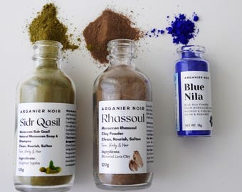 May include: Three glass bottles of Arganier Noir products: Sidr Qasil shampoo, Rhassoul clay powder, and Blue Nila powder. The Sidr Qasil bottle contains a green powder, the Rhassoul bottle contains a brown powder, and the Blue Nila bottle contains a blue powder.