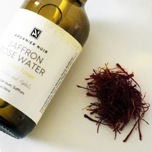 May include: A glass bottle of Arganier Noir Saffron Rose Water mist toner. The bottle is labeled with the product name and ingredients. A small pile of dried saffron threads is visible in the background.
