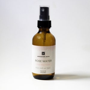 Puede incluir: Una botella de vidrio transparente con una boquilla de pulverización negra llena de agua de rosas. La botella tiene una etiqueta blanca con el texto "ARGANIER NOIR ROSE WATER Face Mist Toner Refresh, Soothe and Hydrate Eau de rose 2 fl oz 59ml".