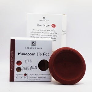 Puede incluir: Un tarro de terracota de tinte para labios y mejillas marroquí. El tarro está etiquetado como "Arganier Noir Moroccan Lip Pot Lip & Cheek Stain". Los ingredientes se enumeran como "Clay Pot, Poppy Petals & Pomegranate" y el producto se describe como "Long Lasting & Buildable Color".