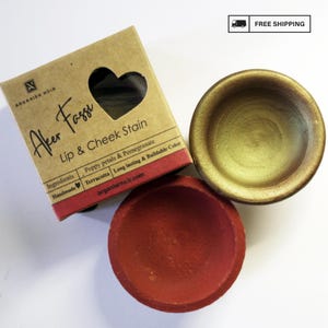 Puede incluir: Vista superior de un producto de tinte para labios y mejillas. El producto incluye una caja de cartón con el texto "Aker Fassi Lip & Cheek Stain" y dos pequeños cuencos de terracota, uno rojo y otro dorado.