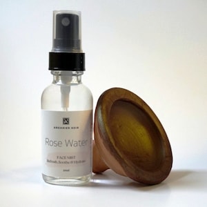 Puede incluir: Una botella de vidrio transparente con un pulverizador negro y una etiqueta blanca que dice "Rose Water Face Mist Refresh, Soothe & Hydrate 20ml" y un pequeño logotipo negro con el texto "Arganier Noir". La botella está al lado de un cuenco de cerámica marrón.