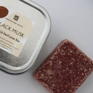 May include: A silver tin with a white label that reads "Arganier Noir Black Musk Solid Perfume Bar". A brown solid perfume bar is next to the tin.