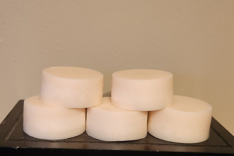 Puede incluir: Una pila de seis jabones redondos de color blanquecino. Las barras cil&iacute;ndricas tienen un color suave y cremoso y est&aacute;n dispuestas en un patr&oacute;n ligeramente escalonado. Los jabones est&aacute;n sobre una superficie oscura contra un fondo neutro.