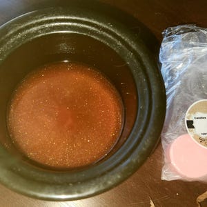 Puede incluir: Un recipiente de cer&aacute;mica negro lleno de un l&iacute;quido &aacute;mbar brillante. A la derecha, una bolsa de pl&aacute;stico transparente contiene varias ceras perfumadas rosas y circulares. La bolsa tiene una etiqueta que dice "Candles by Jess Scac" y "Strawberry Shortcake."