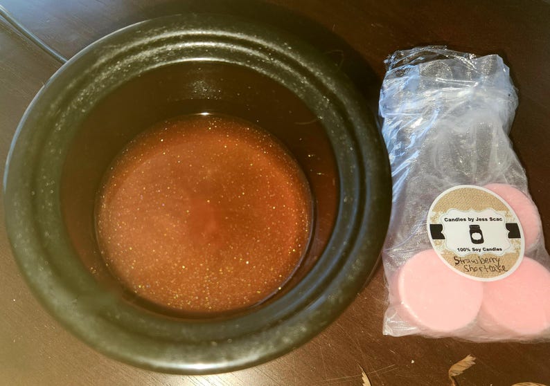 Puede incluir: Un recipiente de cer&aacute;mica negro lleno de un l&iacute;quido &aacute;mbar brillante. A la derecha, una bolsa de pl&aacute;stico transparente contiene varias ceras perfumadas rosas y circulares. La bolsa tiene una etiqueta que dice "Candles by Jess Scac" y "Strawberry Shortcake."