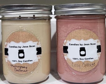 May include: Two glass jar candles with silver lids. One is beige and labeled "Mahogany Coconut." The other is pink and labeled "Cozy Cottage." Both have a burlap-style label with "Candles by Jess Scac" and "100% Soy Candles."