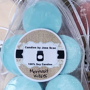 Puede incluir: Primer plano de varias velas redondas de cera de soja azul claro en una bolsa blanca transparente. Las velas est&aacute;n etiquetadas como "Mermaid Kisses" y "Candles by Jess Scac."