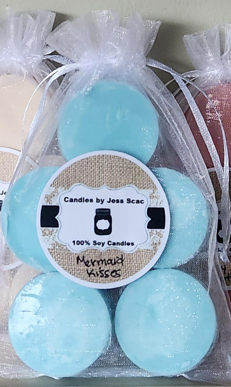Puede incluir: Primer plano de varias velas redondas de cera de soja azul claro en una bolsa blanca transparente. Las velas est&aacute;n etiquetadas como "Mermaid Kisses" y "Candles by Jess Scac."