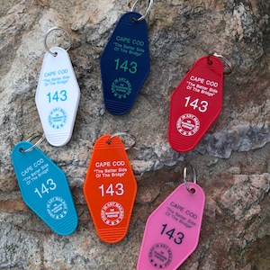 Puede incluir: Seis llaveros coloridos con forma de llaves de habitación de motel. Cada llavero tiene el texto "Cape Cod" y "The Better Side Of The Bridge" con el número "143" en cada uno. Los llaveros son azules, rojos, naranjas, rosas, blancos y azul oscuro.