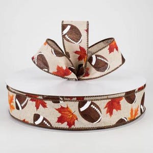 May include: A roll of brown and beige ribbon with a pattern of footballs and autumn leaves. The ribbon is on a white spool.
