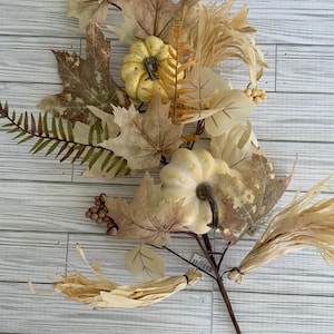 May include: A fall-themed decorative stem featuring faux pumpkins, leaves, and dried grasses. The pumpkins are cream-colored with dark brown stems. The leaves are various shades of brown and tan. The stem is ideal for autumn home decor.