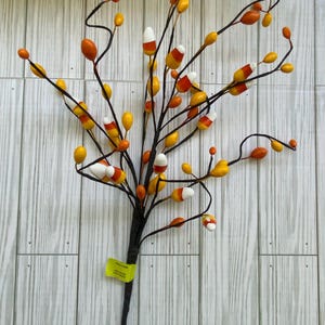 May include: A decorative Halloween-themed branch with black stems and various orange, yellow, and white candy corn-shaped ornaments. The branch is set against a light wood-grain background, creating a festive autumn display.