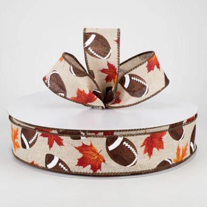 May include: A roll of brown and beige ribbon with a pattern of footballs and autumn leaves. The ribbon is on a white spool.