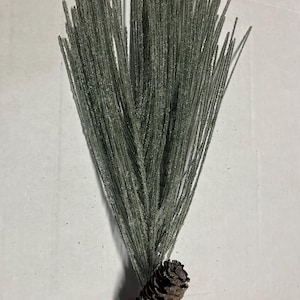 May include: A decorative artificial pine branch with a brown pine cone at the base. The green needles are dusted with white glitter. The pine cone is brown with dark brown accents. The stem is black.