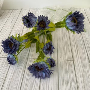 May include: A bouquet of artificial blue sunflowers with green leaves. The sunflowers are arranged in a fan shape and have a realistic appearance.