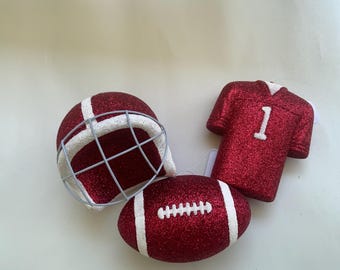 May include: Red glittery football-themed ornaments, including a helmet, jersey with the number 1, and a football. The ornaments have white accents and are likely for holiday or sports-themed decor.