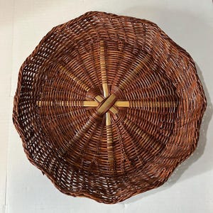 May include: A round, shallow basket with a scalloped edge, woven from dark brown reed or willow. The center features a star-shaped pattern created by lighter, natural-colored bamboo or wood spokes radiating outward from the middle point.