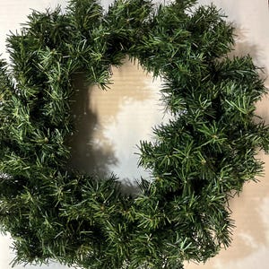 May include: A circular artificial wreath constructed from dense, dark green plastic pine needles. The foliage is arranged in a thick, full ring pattern, creating a textured appearance with multiple individual needle-like branches extending outward.