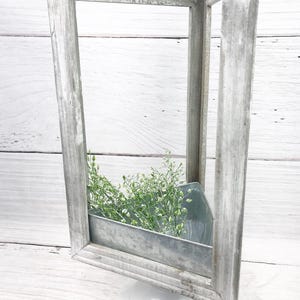 May include: A distressed, rectangular, gray wooden frame with a galvanized metal planter box. The planter holds green sprigs and sits on a white pedestal. The frame is weathered, giving it a rustic appearance.