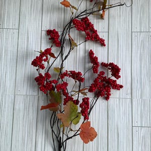 May include: A decorative artificial berry stem with clusters of bright red berries and a few autumn-colored leaves. The berries are attached to dark, slender branches. The stem is displayed against a light gray and white wood-grain background.