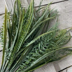 May include: A bundle of artificial greenery, featuring various shades of green. The arrangement includes long, slender blades of grass, fern-like sprigs, and other leafy textures. The faux plants are arranged on a weathered wooden surface.