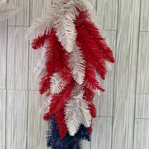 May include: A red, white, and blue artificial Christmas tree garland. The garland is made of plastic and is hanging from a white hook. The garland is perfect for decorating for the holidays.