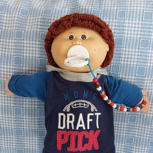 May include: A Cabbage Patch Kid doll wearing a brown knitted hat, a blue and white shirt with the text "MOM'S DRAFT PICK", grey pants, and white sneakers. The doll is holding a white pacifier with a blue and red beaded chain.