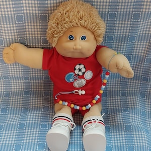 Puede incluir: Una muñeca Cabbage Patch Kid con cabello rubio, ojos azules y una camiseta roja con un diseño deportivo. La muñeca lleva un collar de cuentas y zapatos blancos.