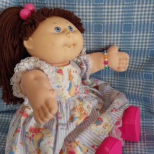 Puede incluir: Una muñeca vintage con cabello castaño en cola de caballo, ojos azules y expresión sonriente. La muñeca viste un vestido floral con estampado de Peter Rabbit y zapatos rosas. Una pulsera de cuentas coloridas adorna su muñeca. La muñeca está sentada sobre una superficie a cuadros azul y blanco.