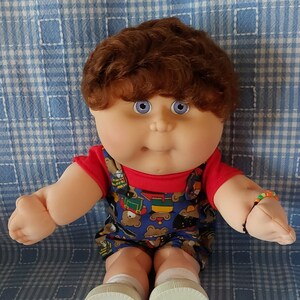 K&ouml;nnte beinhalten: Eine Cabbage Patch Kid Puppe mit braunen Haaren, blauen Augen, einem roten Shirt und einer blauen und gelben gemusterten Latzhose. Die Puppe tr&auml;gt wei&szlig;e Schuhe und ein Armband am Handgelenk.