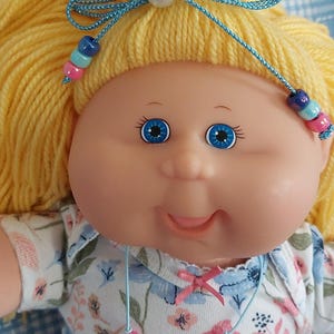 Puede incluir: Muñeca vintage con cabello de hilo amarillo peinado con un lazo azul y adornos de cuentas. La muñeca tiene ojos azules y lleva un body con estampado floral y un collar de cuentas. La muñeca lleva pantalones rosas.