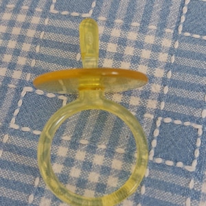 May include: A clear, yellow baby pacifier with a round ring and a flat, circular shield. The pacifier is made of a translucent material and is set against a blue and white checkered background.