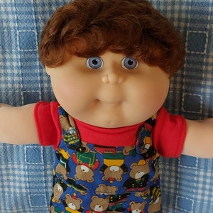 May include: A Cabbage Patch Kid doll with brown hair, blue eyes, and a red shirt. The doll is wearing blue overalls with a pattern of teddy bears. The doll is wearing white shoes.