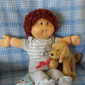 Puede incluir: Una muñeca Cabbage Patch Kid con cabello rizado marrón que lleva un mono a rayas gris y blanco con un lazo rojo y pantalones grises. La muñeca sostiene un perro de peluche marrón y está sentada sobre una manta azul y blanca con un patrón de perro y pelota.