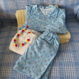 Pode incluir: Um conjunto de roupa de bebé com uma parte de cima e calças azuis claras com um padrão de peixes, uma chupeta e uma fralda. O conjunto está colocado sobre uma manta de malha amarela, tudo sobre uma superfície azul estampada.