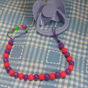 May include: A lavender pacifier with a beaded chain. The chain features purple, pink, and green beads spelling out "KPG". The pacifier has a flower design on the front.