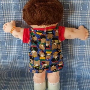 K&ouml;nnte beinhalten: Eine Cabbage Patch Kid Puppe mit braunen Haaren, die ein blaues und rotes Outfit mit einem B&auml;renmuster tr&auml;gt. Die Puppe tr&auml;gt wei&szlig;e Schuhe und sitzt auf einer blau-wei&szlig; karierten Decke.