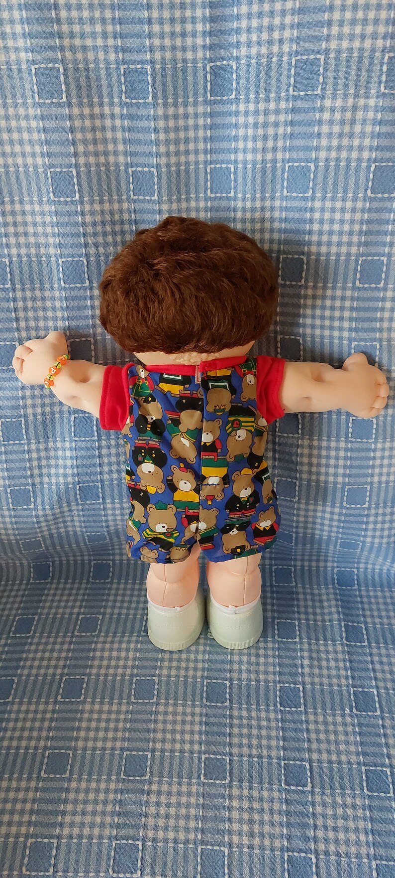 K&ouml;nnte beinhalten: Eine Cabbage Patch Kid Puppe mit braunen Haaren, die ein blaues und rotes Outfit mit einem B&auml;renmuster tr&auml;gt. Die Puppe tr&auml;gt wei&szlig;e Schuhe und sitzt auf einer blau-wei&szlig; karierten Decke.