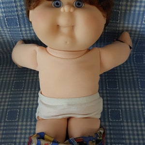 K&ouml;nnte beinhalten: Eine Cabbage Patch Kid Puppe mit braunen Haaren, blauen Augen und einer wei&szlig;en Windel. Die Puppe tr&auml;gt ein rot-blaues gemustertes Hemd mit rotem Kragen und wei&szlig;en Turnschuhen.
