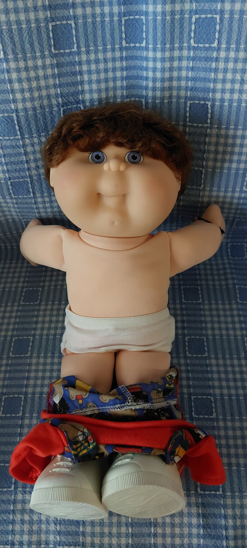 K&ouml;nnte beinhalten: Eine Cabbage Patch Kid Puppe mit braunen Haaren, blauen Augen und einer wei&szlig;en Windel. Die Puppe tr&auml;gt ein rot-blaues gemustertes Hemd mit rotem Kragen und wei&szlig;en Turnschuhen.
